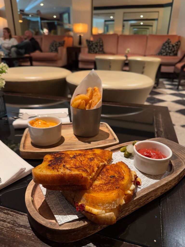 Toasted sandwich served with thick chips and dipping sauces on wooden boards in a softly lit hotel lounge with pink sofas.