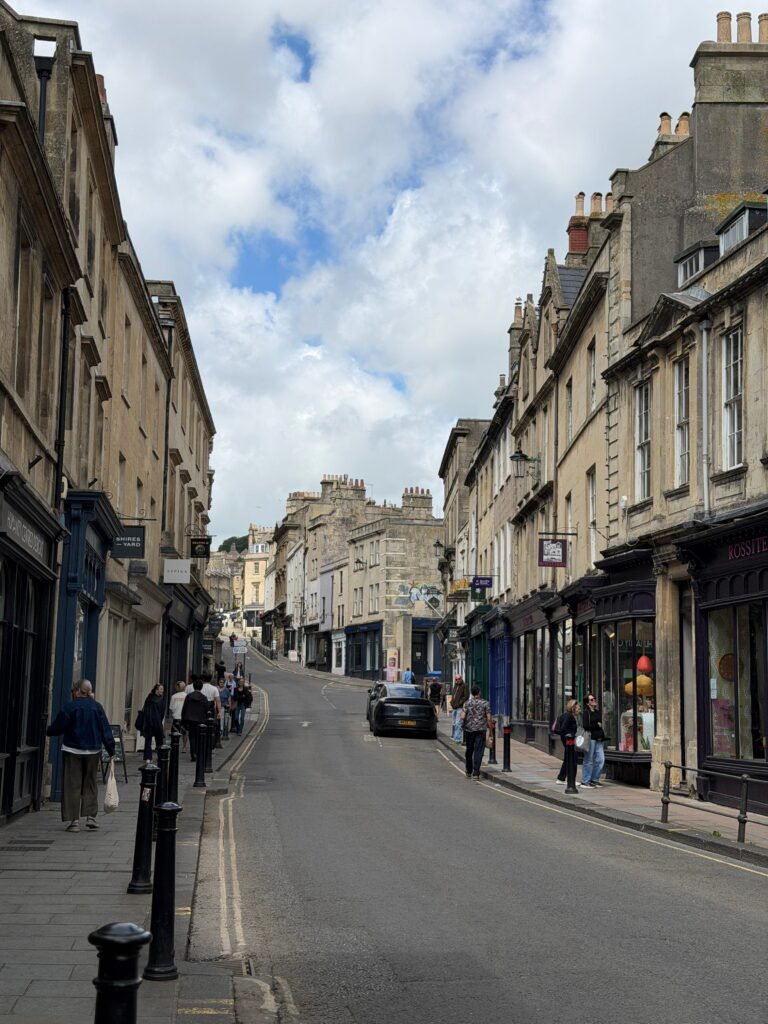 A gently sloping street in Bath lined with historic honey coloured Georgian stone buildings, independent shops and cafés. Pedestrians walk along the pavements beneath a partly cloudy sky, with black bollards and traditional shopfronts adding to the character of the city centre.