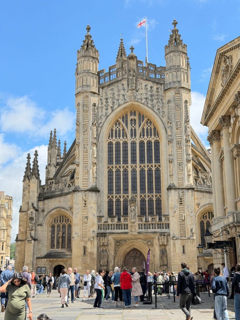 The ornate Gothic façade of Bath Abbey with its enormous stained glass window and two intricately carved stone towers. Visitors gather in the square outside beneath a bright blue sky.