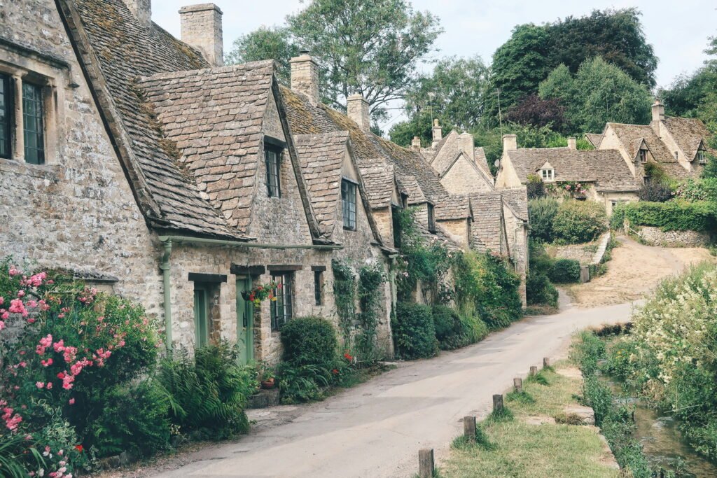 Traditional honey coloured stone cottages with steep tiled roofs line a narrow lane beside a small stream. Pink flowers and lush cottage gardens soften the historic village scene.