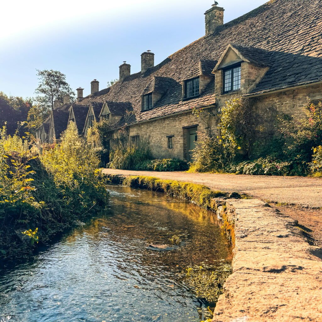 Honey coloured stone cottages with steep tiled roofs and dormer windows beside a shallow stream. Warm sunlight falls across the cottages and waterside plants.