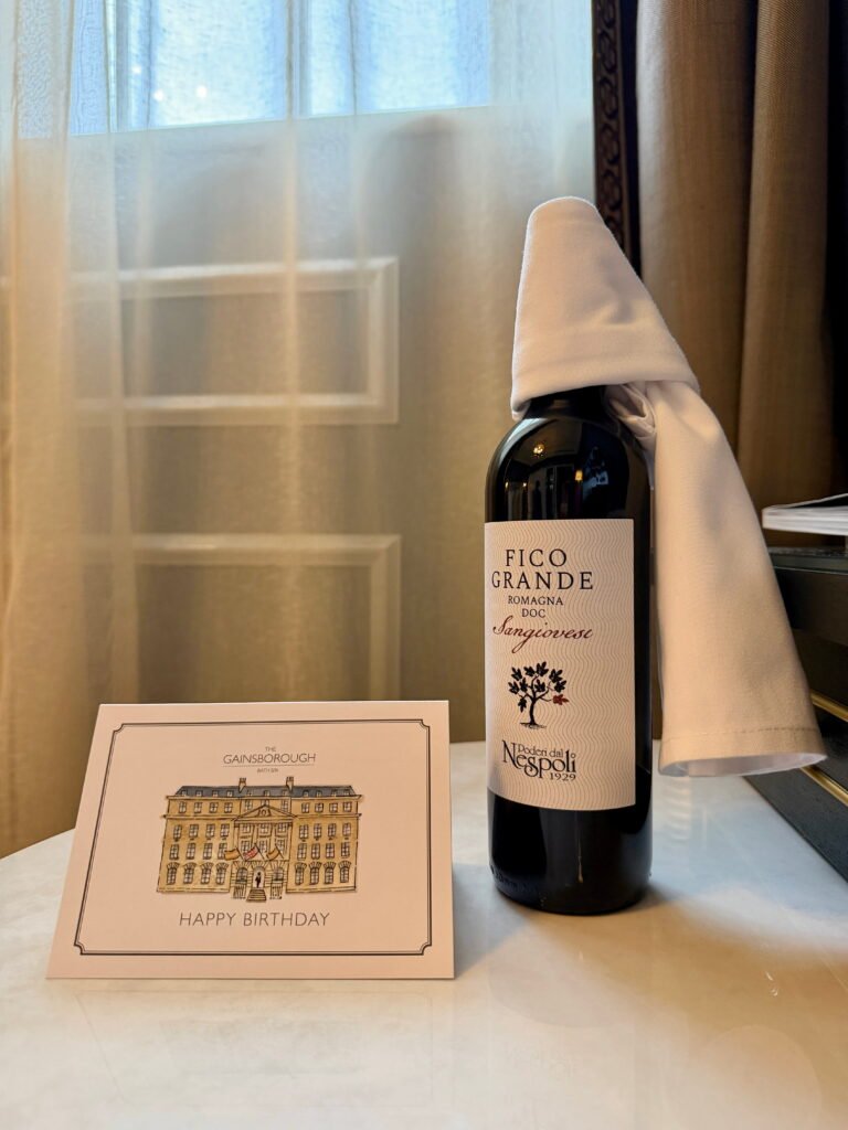 A bottle of Fico Grande Sangiovese red wine with a white napkin draped over the top sits beside a birthday card from The Gainsborough Bath Spa. The card features an illustration of the hotel building and the words "Happy Birthday".