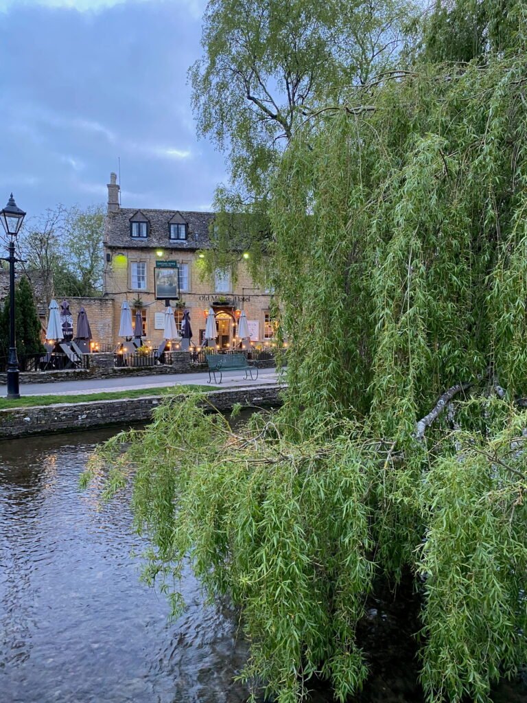 The Old Manse Hotel glowing with warm lights beside the river at dusk. A large weeping willow fills the foreground with long green branches hanging over the water.