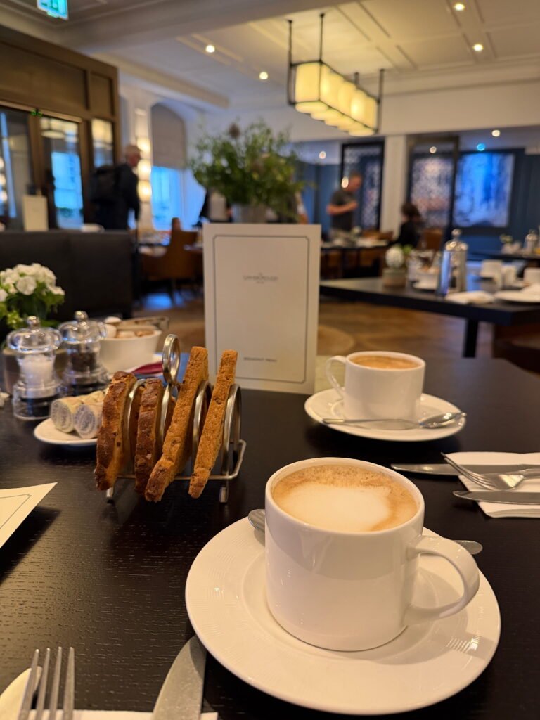 Two white cups of frothy coffee sit on a dark wooden table alongside a silver toast rack filled with slices of toast. The Gainsborough Bath Spa breakfast room is softly blurred in the background with warm pendant lighting and brown leather chairs.