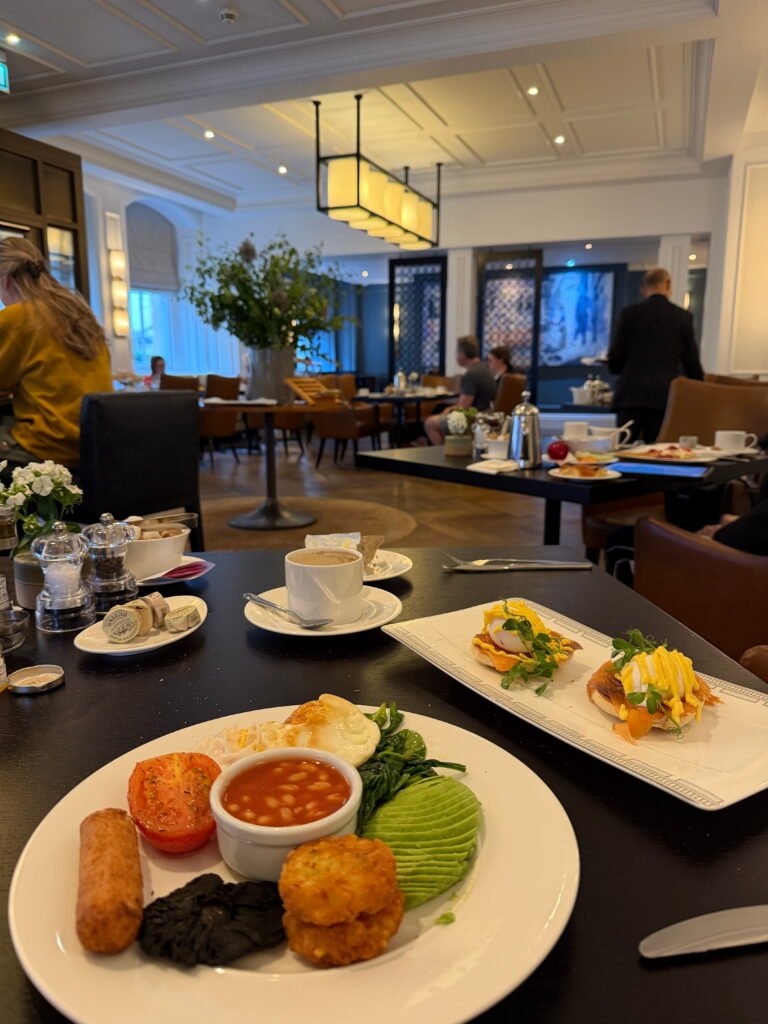 A full English breakfast at The Gainsborough Bath Spa featuring a fried egg, sausage, black pudding, hash browns, baked beans, grilled tomato, spinach and sliced avocado. A separate rectangular plate holds two portions of eggs Benedict with hollandaise sauce and fresh herbs.