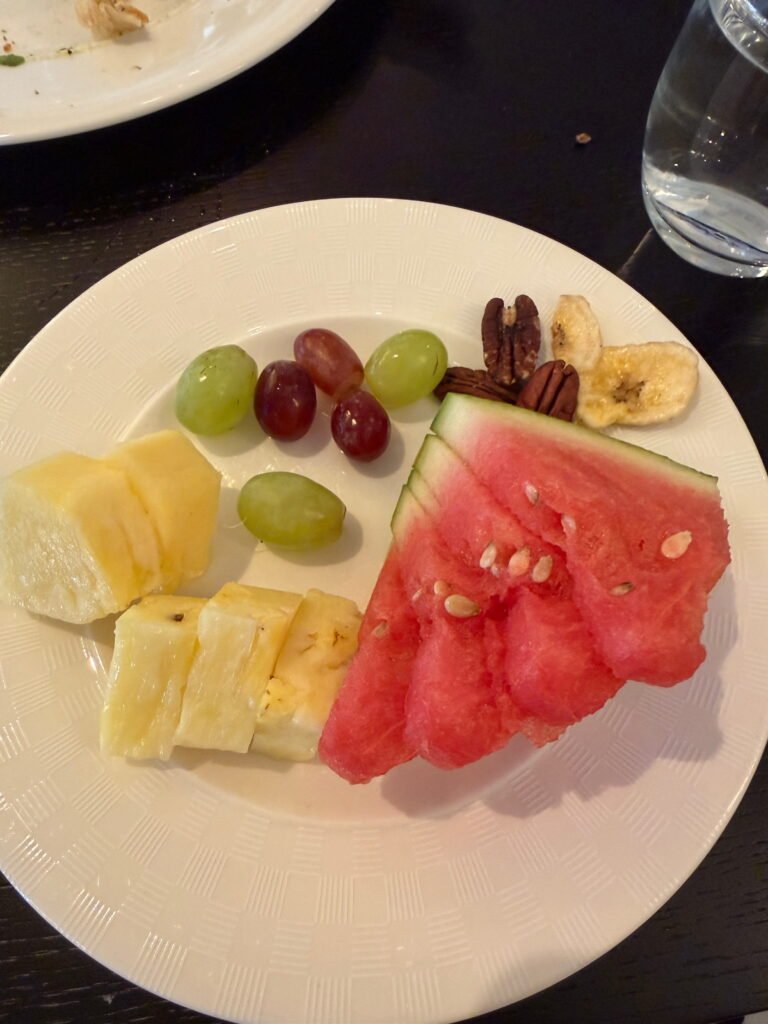 A white plate of fresh fruit featuring slices of watermelon, pineapple, red and green grapes, pecans and dried banana pieces. The colourful fruit is arranged on a white patterned plate on the breakfast table at The Gainsborough Bath Spa.