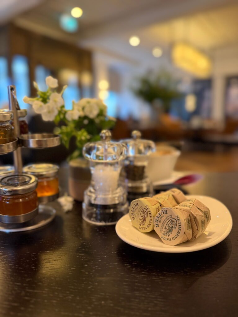 Individually wrapped Netherend Farm butter portions sit on a white plate beside glass salt and pepper mills and small jars of preserves. A small arrangement of white flowers decorates the breakfast table at The Gainsborough Bath Spa.
