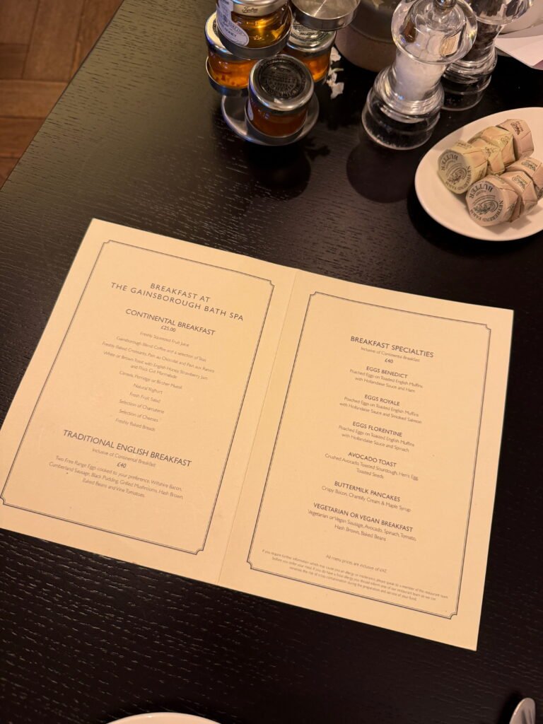 An open breakfast menu from The Gainsborough Bath Spa rests on a dark wooden table beside small jars of preserves and individually wrapped butter. The menu offers continental and traditional English breakfasts alongside eggs Benedict, eggs Royale, eggs Florentine, avocado toast, buttermilk pancakes and vegetarian or vegan breakfast options.