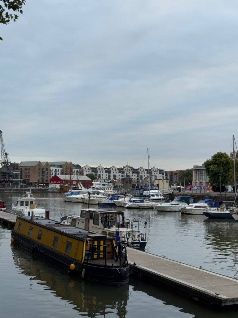 Boats and narrowboats moored around a Bristol harbour beneath an overcast sky. Waterfront buildings and an old industrial crane line the opposite side.