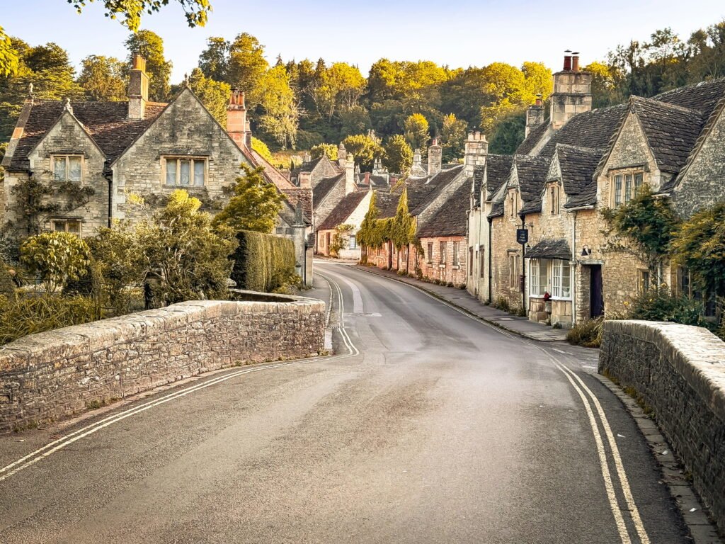 A quiet road curves across a stone bridge and between traditional honey coloured cottages. Late afternoon sunlight gives the trees and historic village buildings a warm golden glow.