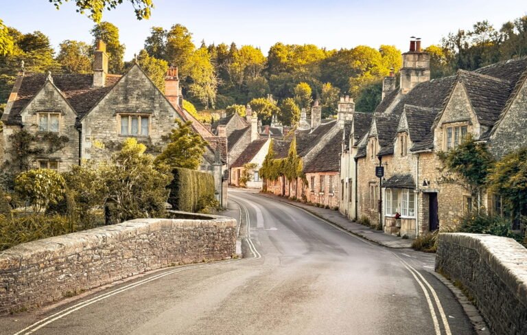 A quiet road curves across a stone bridge and between traditional honey coloured cottages. Late afternoon sunlight gives the trees and historic village buildings a warm golden glow.