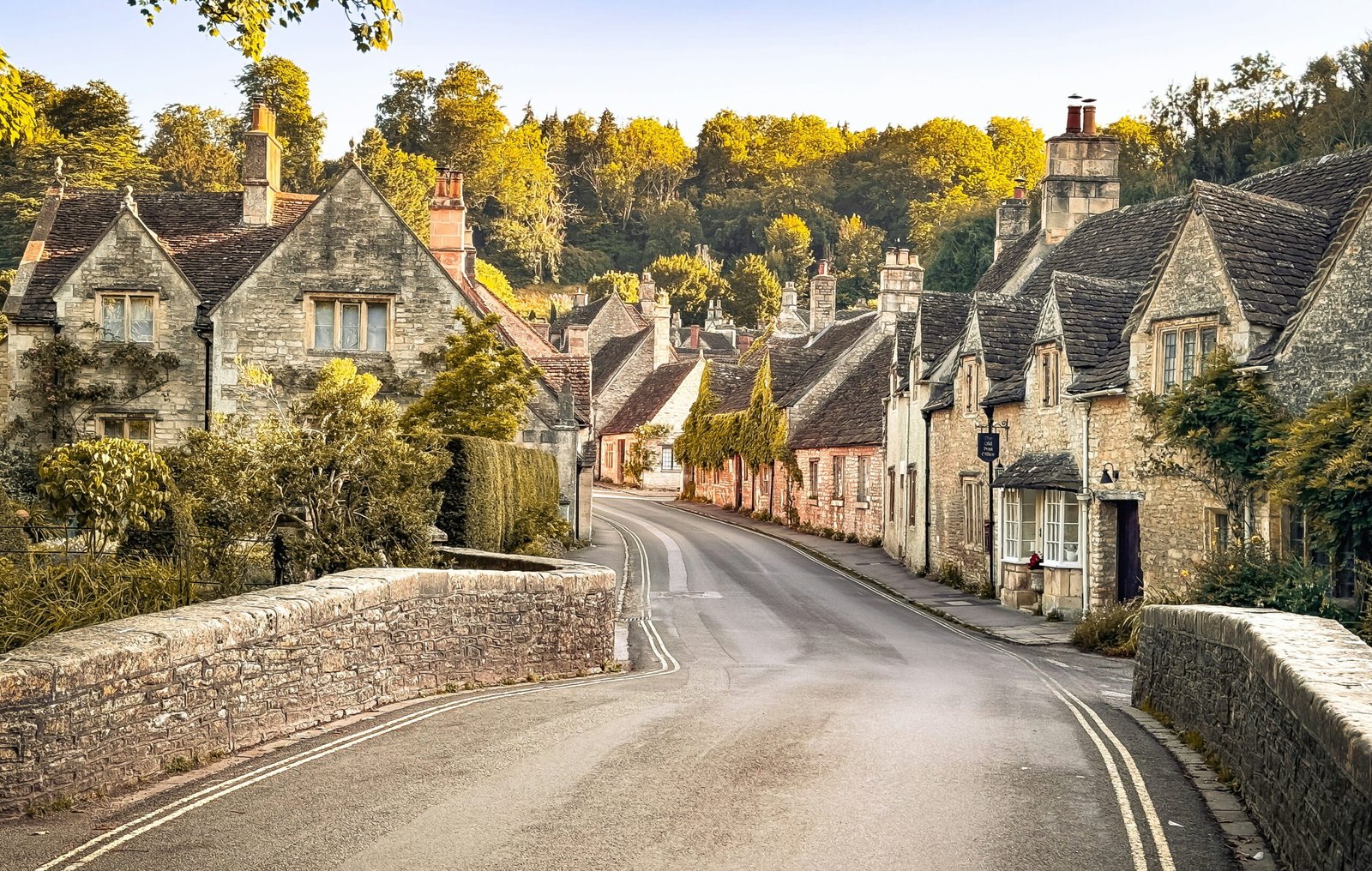 A quiet road curves across a stone bridge and between traditional honey coloured cottages. Late afternoon sunlight gives the trees and historic village buildings a warm golden glow.