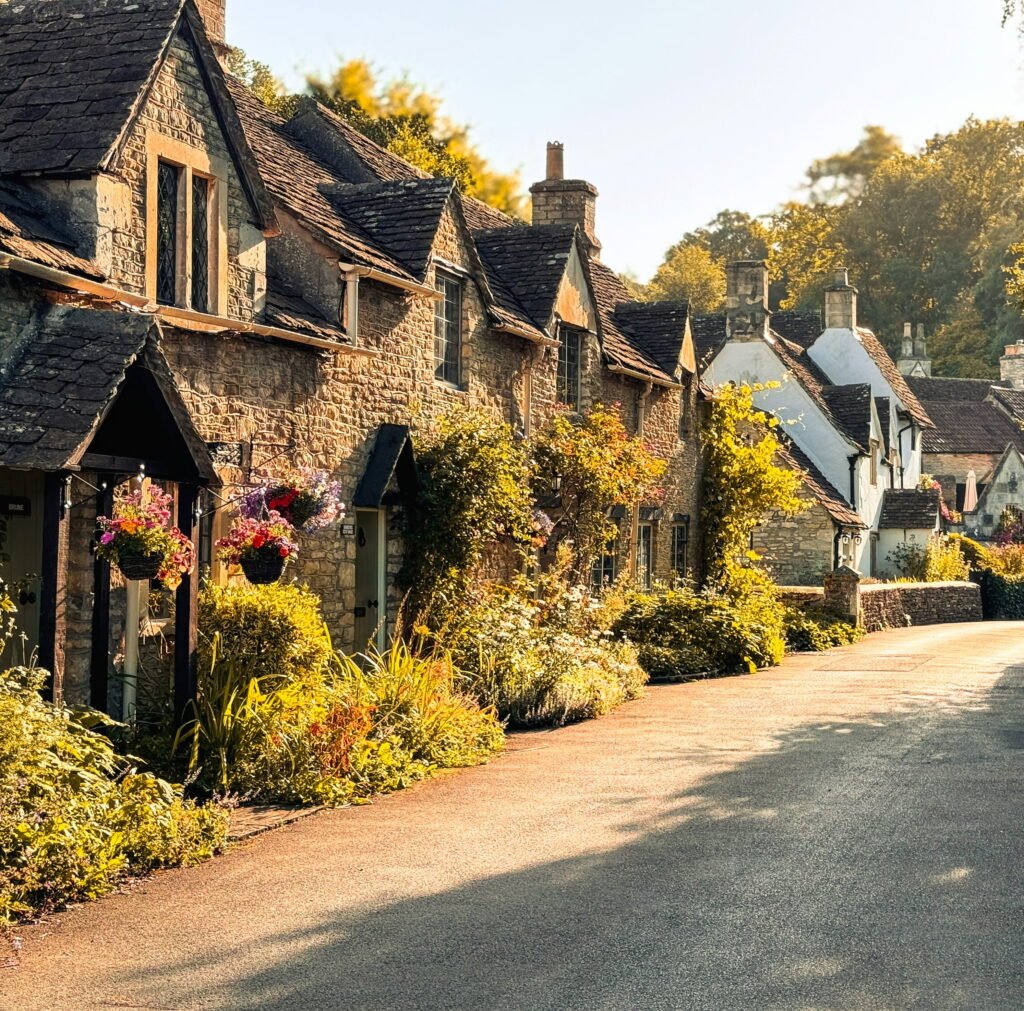 A peaceful village street lined with honey coloured stone cottages and colourful hanging flower baskets. Golden sunlight illuminates the cottage gardens and textured stone walls.