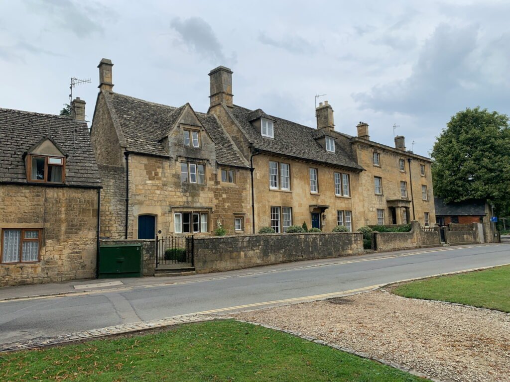 Row of historic honey colored stone houses with steep roofs and tall chimneys facing a quiet road in the Cotswolds. Low stone walls and small front gardens separate the traditional homes from the street.