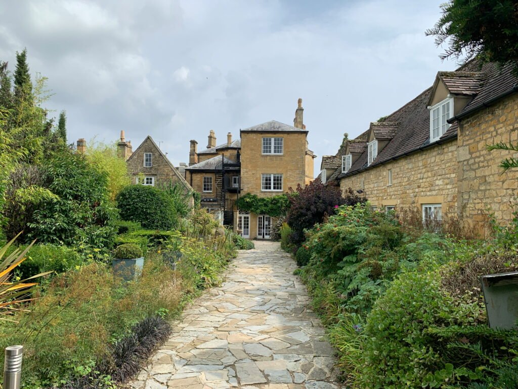Stone pathway leading through a lush garden surrounded by traditional Cotswold stone buildings. Dense shrubs flowers and greenery border the path with historic cottages and chimneys beyond.