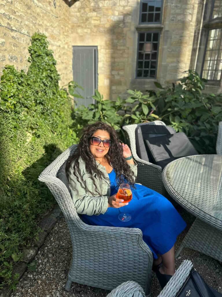 Nishi relaxes in a wicker chair on a sunny garden terrace while holding an Aperol Spritz. She wears sunglasses and a bright blue dress beside tall green hedges and a stone building.