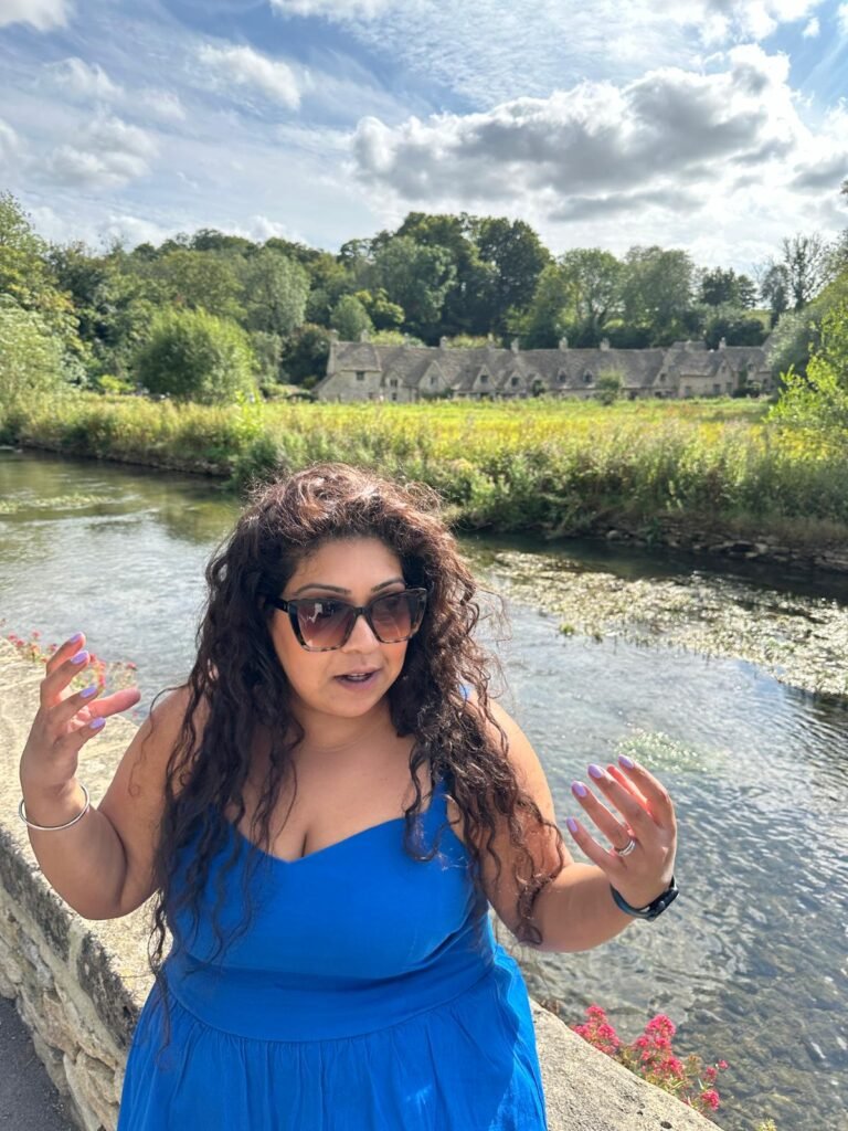 Nishi stands beside a river wearing sunglasses and a bright blue dress while gesturing with both hands. A long row of traditional stone cottages sits across the water behind her.