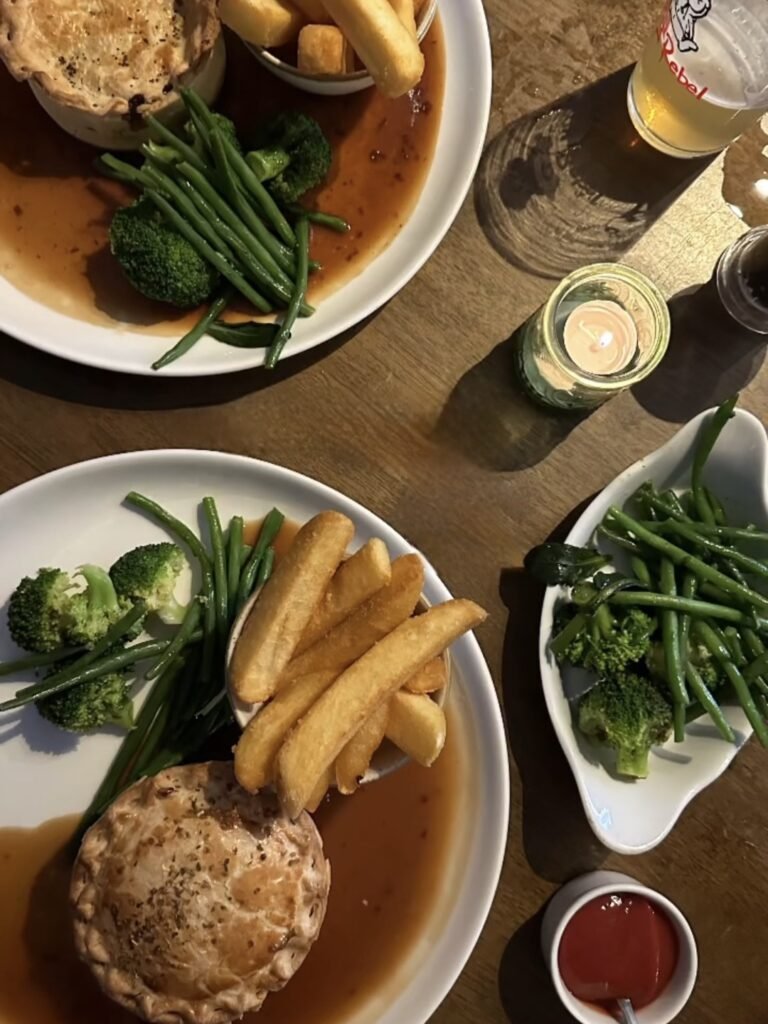 Two plates of pie with thick chips green beans broccoli and gravy sit on a wooden pub table. Extra vegetables ketchup drinks and a lit candle are arranged around the meals.