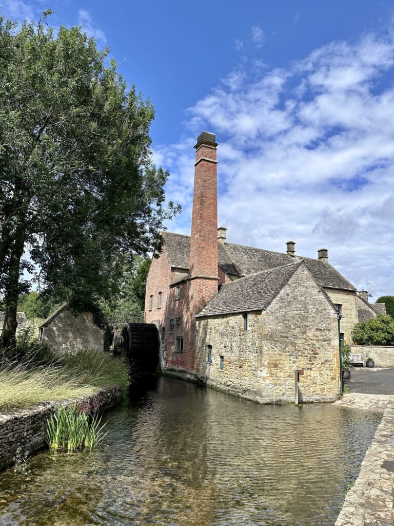 Historic stone and brick mill buildings sit directly beside the water beneath a bright blue sky. A tall red brick chimney and large dark waterwheel rise beside the river.