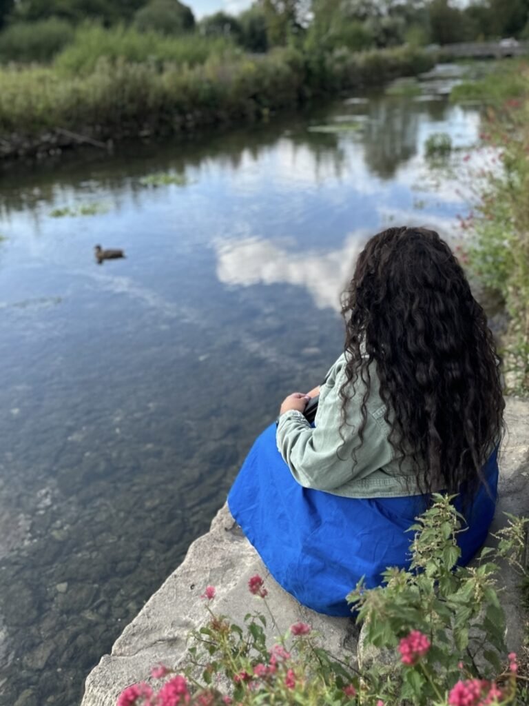 Nishi sits on a stone riverbank with her back to the camera watching a duck on the calm water. She wears a bright blue dress and pale green jacket with pink flowers growing beside her.