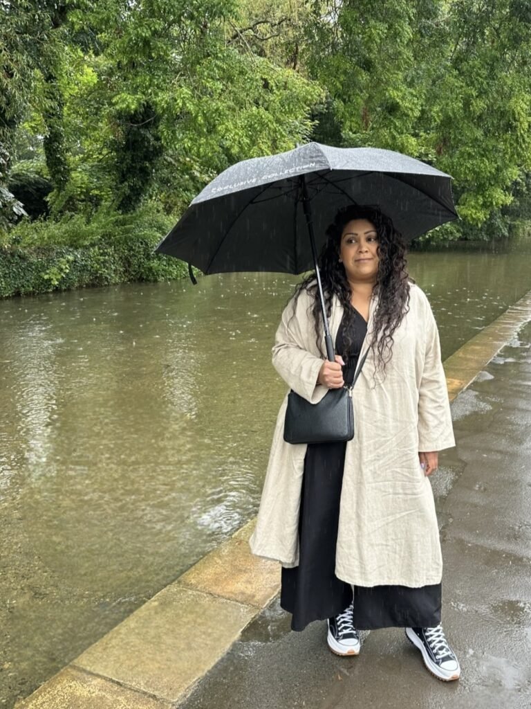 Nishi stands under a black umbrella beside a river during heavy rain. She wears a long cream coat over black clothing while raindrops ripple across the water behind her.