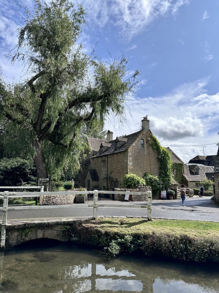 A small stone bridge crosses a stream beneath a large weeping willow in a traditional Cotswold village. Honey coloured cottages and a few people walking through the village appear beyond the bridge.