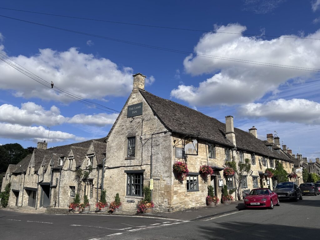 The Lamb Inn occupies a long honey coloured stone building decorated with colourful hanging flower baskets. The words "The Lamb Inn" appear on signs mounted on the historic building beneath a vivid blue and cloudy sky.