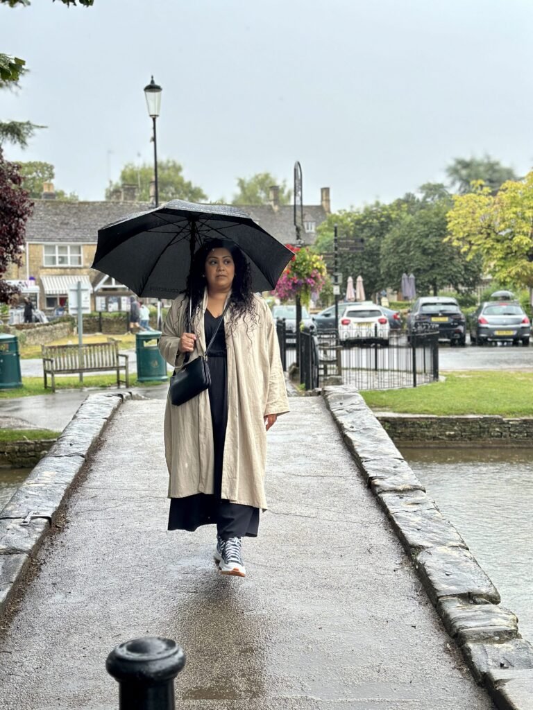 Nishi walks across a wet stone bridge holding a black umbrella in the rain. She wears a long cream coat over black clothing with the village green and stone cottages visible behind her.