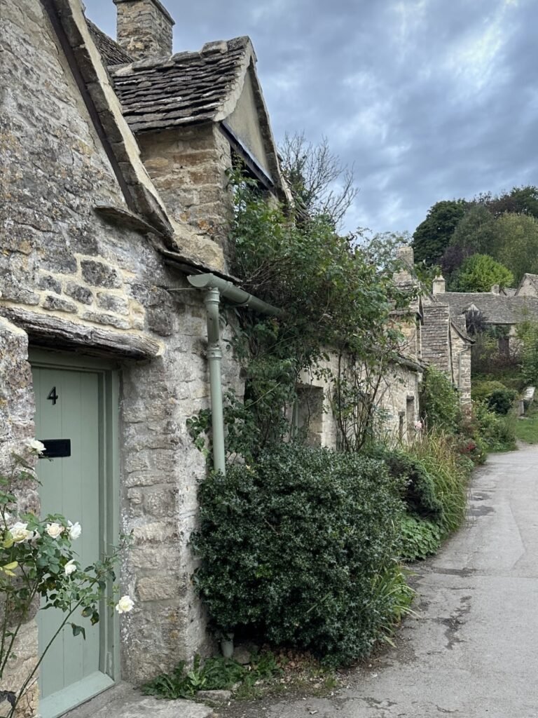 Stone cottage with a pale green door marked "4" beside a narrow village lane. Climbing greenery and white roses soften the weathered Cotswold stone walls beneath an overcast sky.