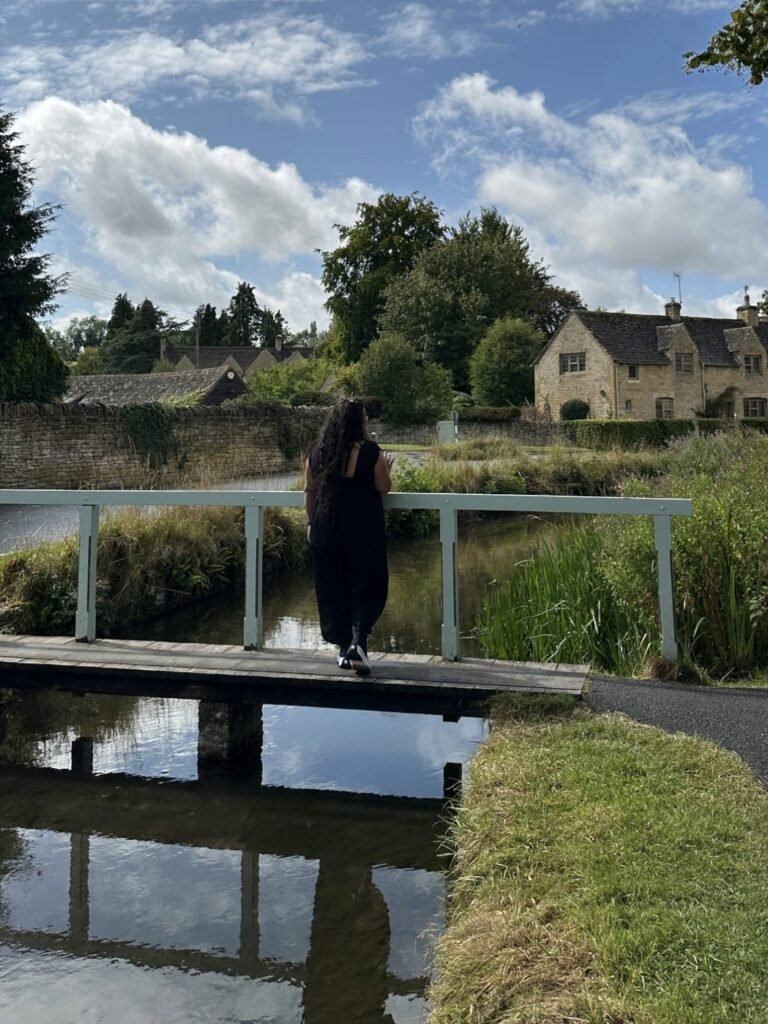 Woman in a black dress standing on a small white footbridge over a stream and looking toward traditional stone cottages. Reeds and trees surround the water beneath a bright blue and cloudy sky.