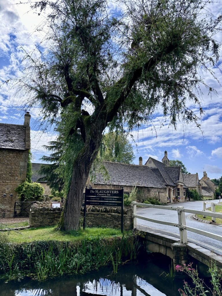 Large willow tree beside a stream and a small white bridge in a Cotswold village. A roadside sign reads "The SLAUGHTERS Country Inn" in front of the stone inn.
