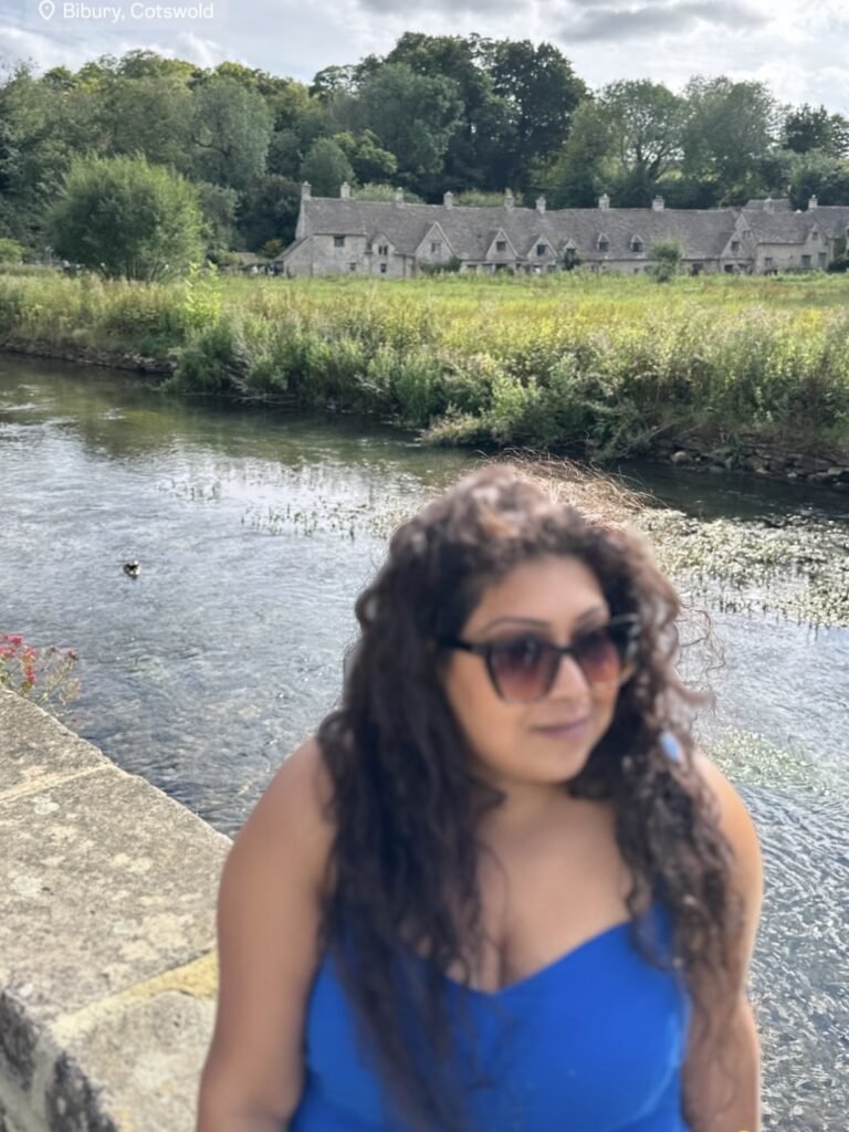 Woman in sunglasses and a bright blue dress beside a river with historic stone cottages across the water. Text at the top reads "Bibury Cotswold."