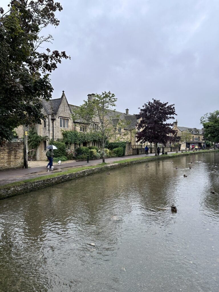 Wide stream running beside historic stone buildings on a rainy day with ducks scattered across the water. People walk beneath umbrellas along the waterside path.