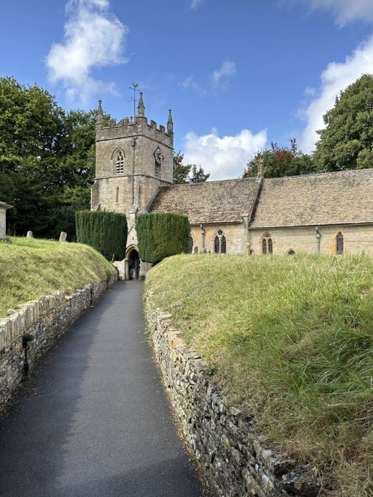 Historic stone church with a square clock tower at the end of a paved path bordered by low stone walls and grassy churchyard. A bright blue sky with white clouds rises above the Cotswold stone building.
