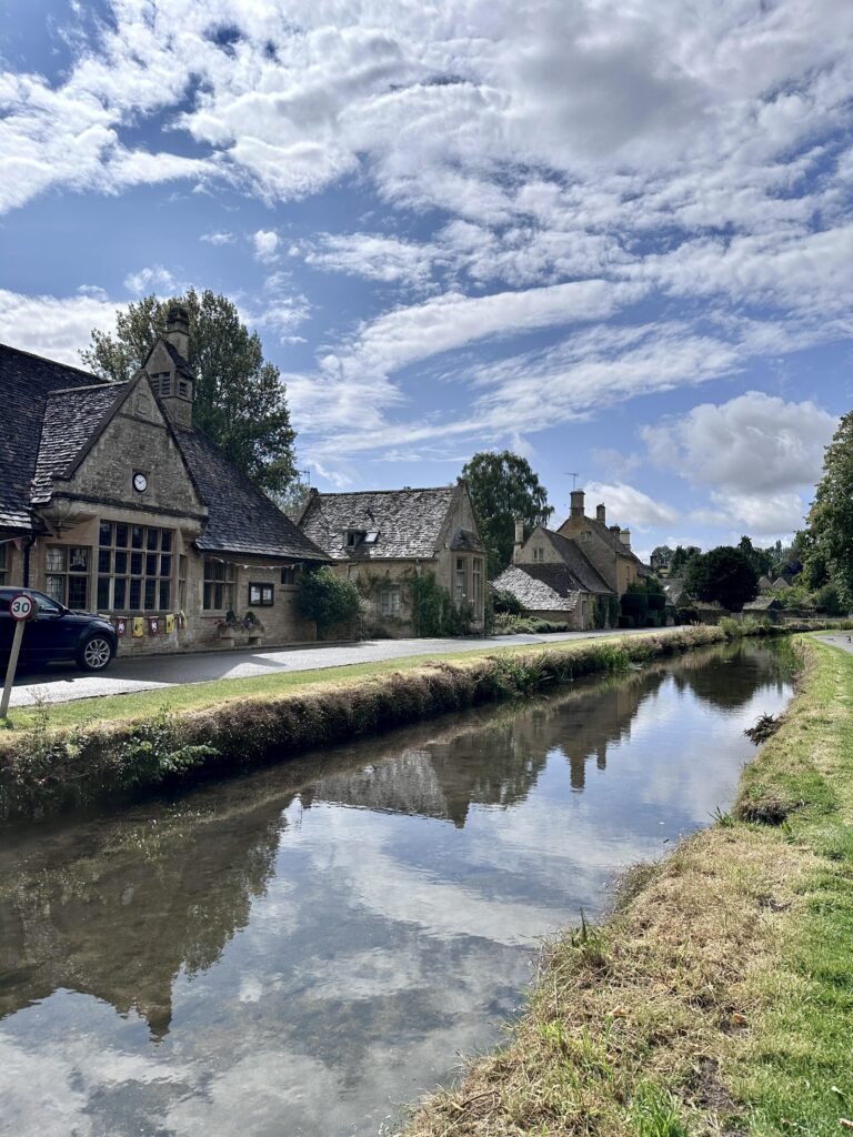 Cotswold stone buildings lining a quiet road beside a clear stream that reflects the cottages and dramatic cloudy sky. A traditional building with a small clock stands prominently beside the water.