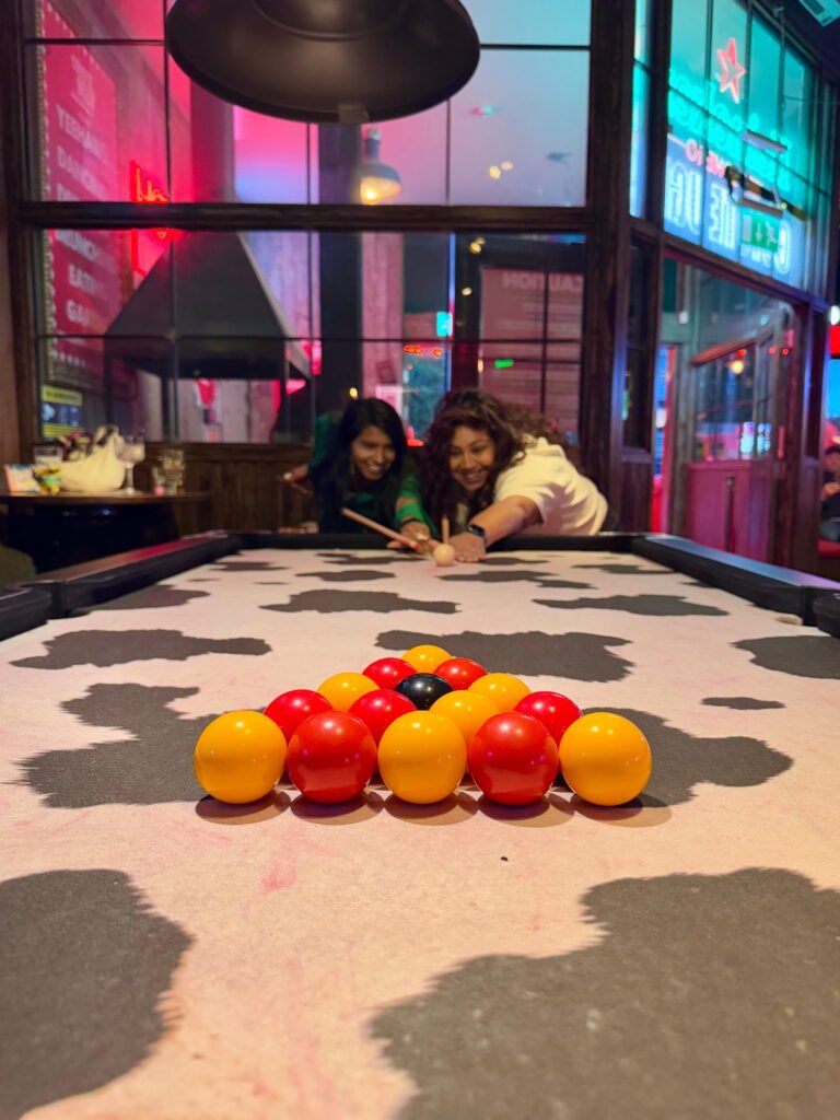 Two women lean across a cow print pool table to take a shot together. Bright red, orange and black balls are racked in the foreground beneath colourful neon lights.
