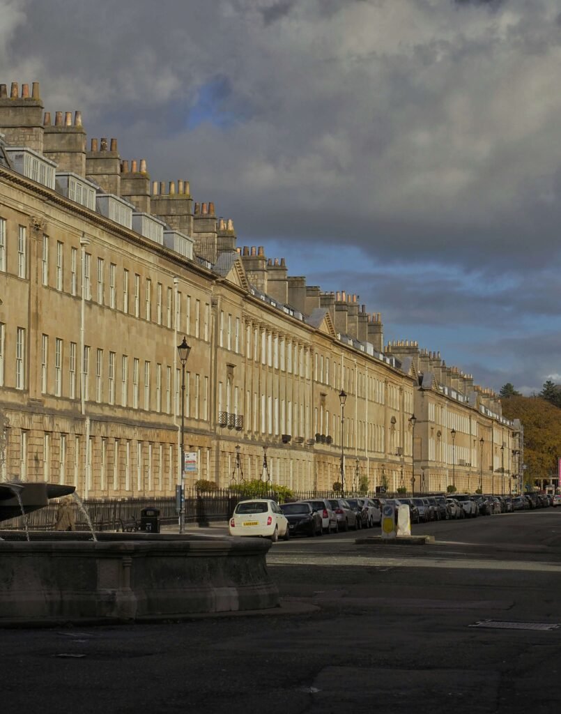 A long row of elegant Georgian townhouses along Great Pulteney Street in Bath, with honey coloured stone façades, tall sash windows and rows of chimney pots stretching into the distance. Golden sunlight illuminates the historic buildings beneath dramatic dark grey clouds, with parked cars, traditional street lamps and part of a stone fountain in the foreground.