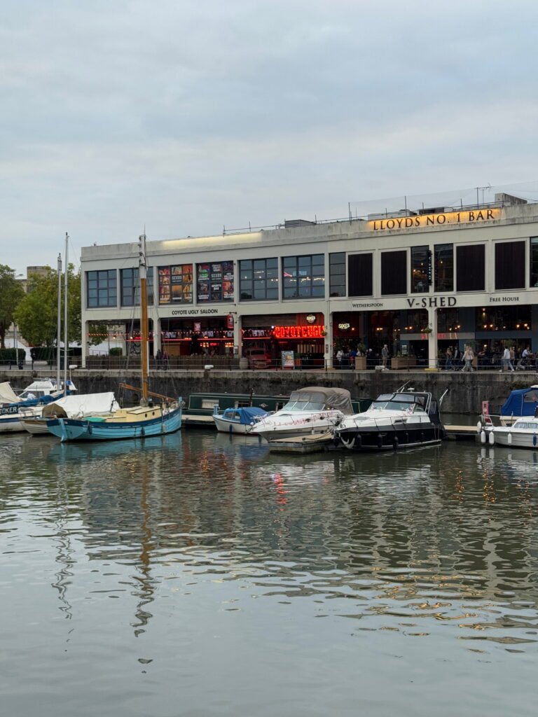 Boats moored in Bristol harbour with the waterfront venues Coyote Ugly Saloon and Lloyds No. 1 Bar behind them. Evening lights reflect softly across the water.