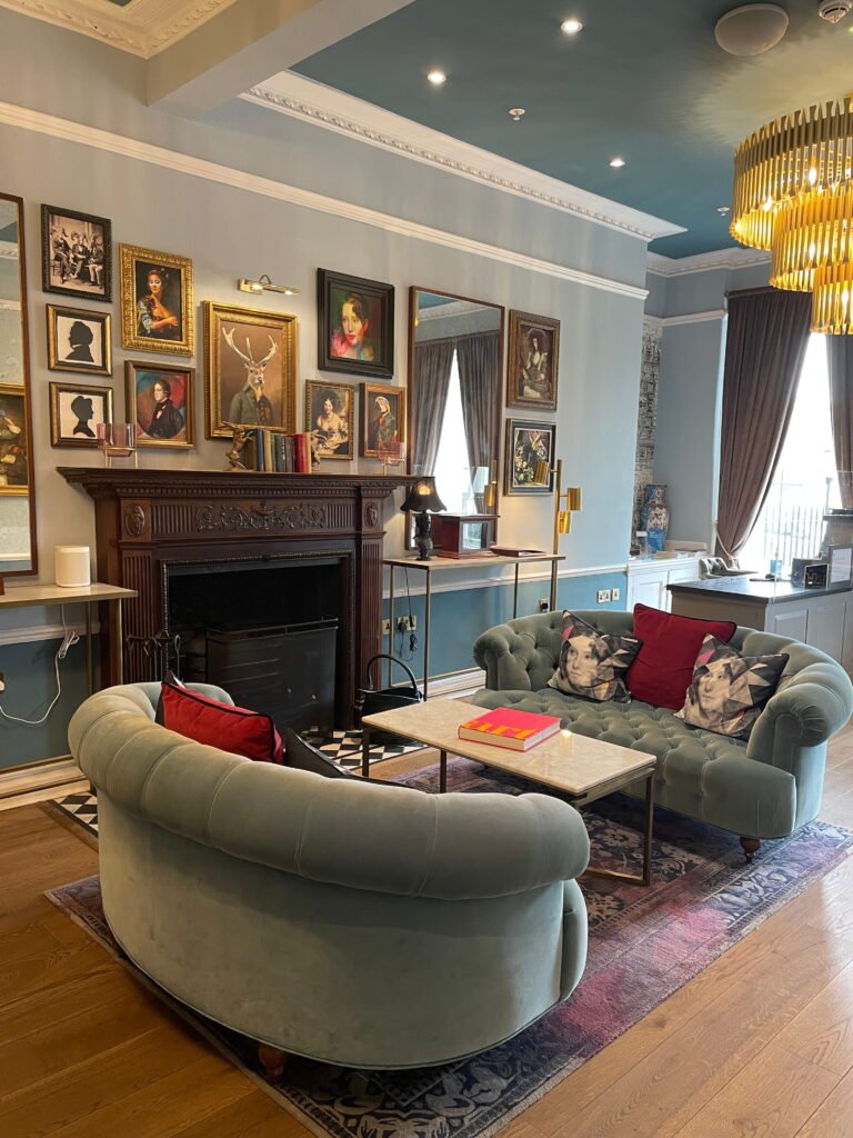 A stylish hotel lounge with two curved sage green velvet sofas surrounding a marble coffee table. An ornate wooden fireplace sits beneath a gallery wall of eclectic framed portraits, including a painting of a stag wearing a suit.