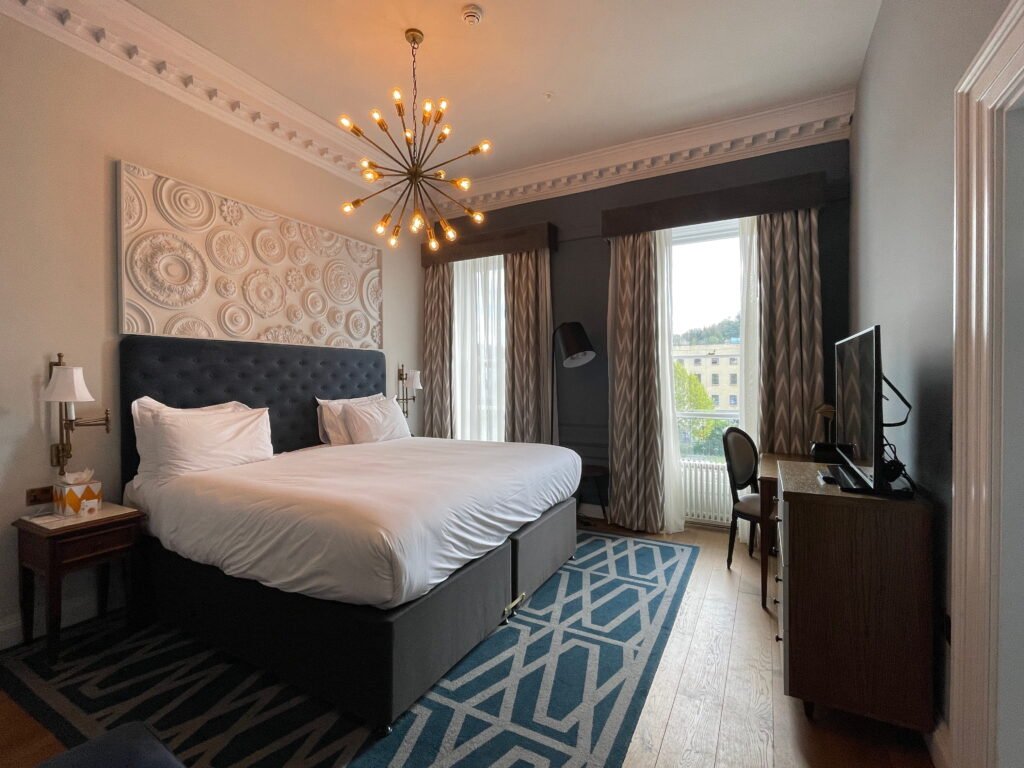 A spacious hotel bedroom with a large double bed dressed in white linen, a dark grey upholstered headboard and a blue geometric rug. A gold starburst chandelier hangs above the bed, while two tall windows overlook Bath's historic buildings.