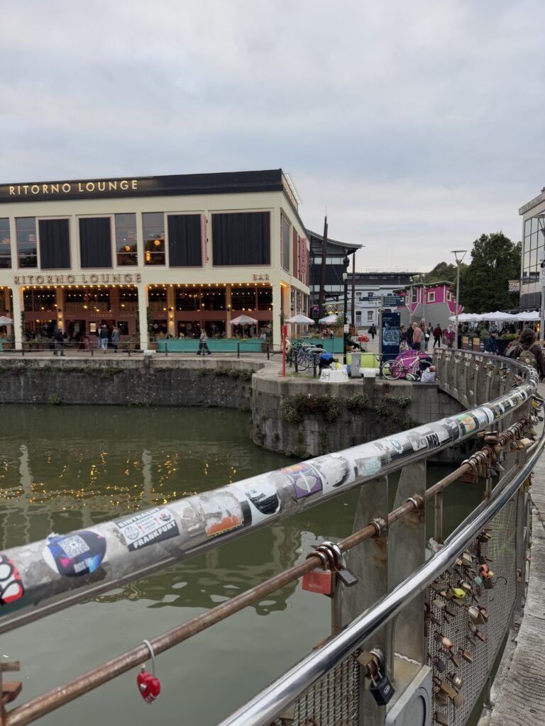 Bristol harbourside view from a bridge covered in padlocks and stickers. Ritorno Lounge glows with warm lights across the water while people walk along the waterfront.