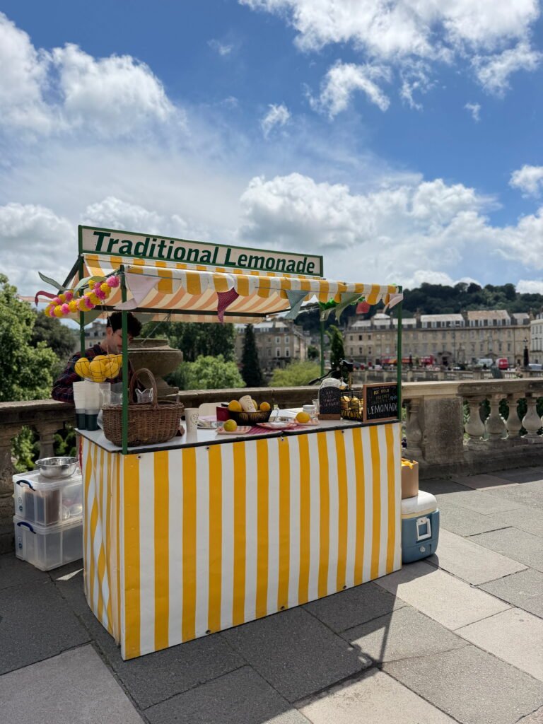 A traditional lemonade stall with yellow and white stripes, colourful bunting and fresh lemons displayed on the counter. The stall sits beside a stone balustrade overlooking Bath and its historic Georgian buildings on a sunny day, with a large sign reading "Traditional Lemonade" above the canopy.