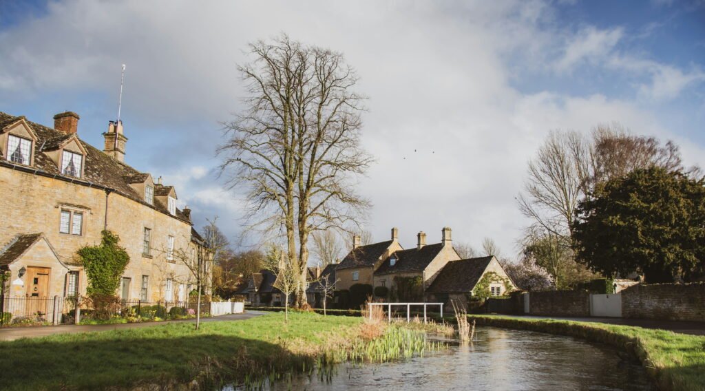 Peaceful Cotswold village scene with honey colored stone cottages surrounding a stream and grassy banks. A small white footbridge crosses the water beneath tall trees and a partly cloudy sky.