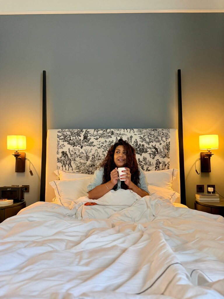 A woman with curly brown hair sits up in a large hotel bed holding a white mug and smiling towards the side of the room. The bed has crisp white bedding and a black and white illustrated headboard with matching bedside lamps casting a warm glow against the grey walls.