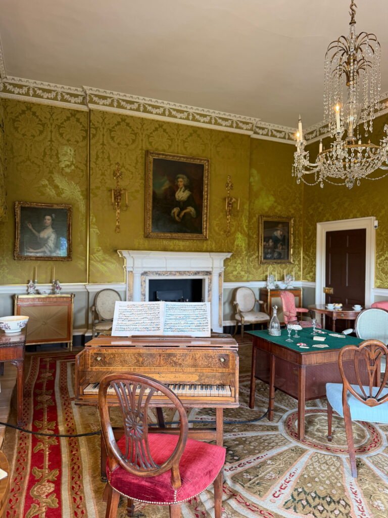 An elegant Georgian drawing room with gold damask wallpaper, a crystal chandelier and antique furniture arranged around a white marble fireplace. An ornate wooden piano with open sheet music sits in the foreground, surrounded by upholstered chairs and framed portraits.