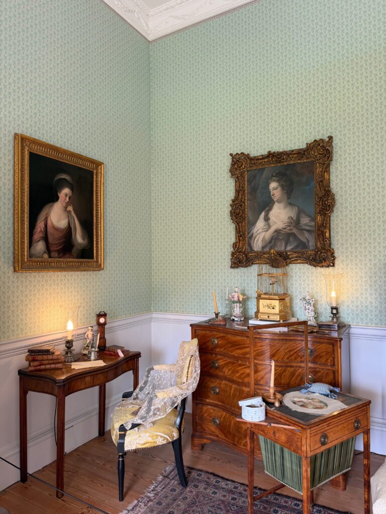 A historic Georgian dressing room with pale green patterned wallpaper, antique wooden furniture and two large portraits of women in ornate gold frames. A lace covered chair sits between a writing desk and a chest of drawers topped with decorative porcelain figurines and a birdcage.