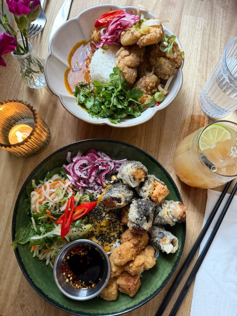 Two colourful Asian inspired dishes served on a wooden restaurant table. One bowl contains white rice, crispy fried pieces, pickled red onion, fresh coriander and sauce, while the other features fried rolls, vegetables, black sesame seeds and a small dish of dipping sauce.