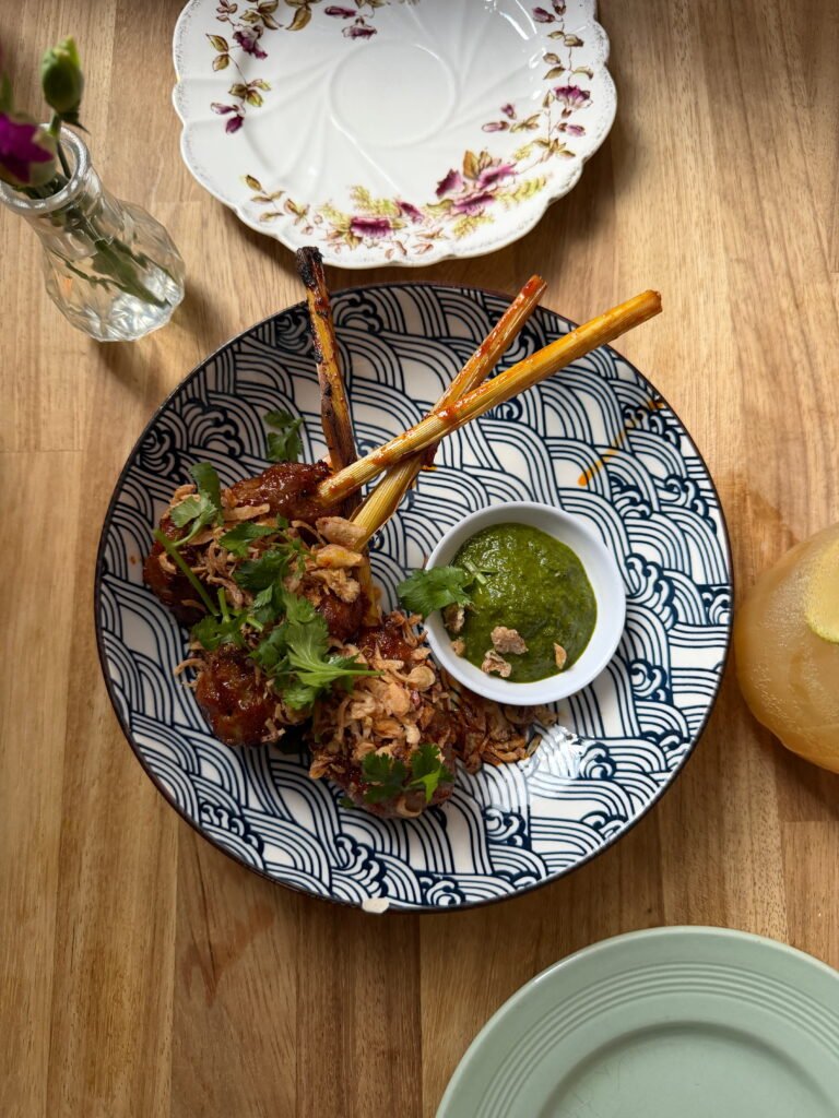Glazed meat on long bones served with fresh coriander and crispy fried onions on a blue and white patterned plate. A small bowl of green dipping sauce sits alongside the meat on the wooden restaurant table.