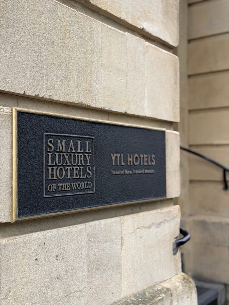 A black and bronze plaque mounted on the pale stone exterior of The Gainsborough Bath Spa displays the words "Small Luxury Hotels of the World" and "YTL Hotels". The YTL Hotels tagline reads "Treasured Places. Treasured Moments".