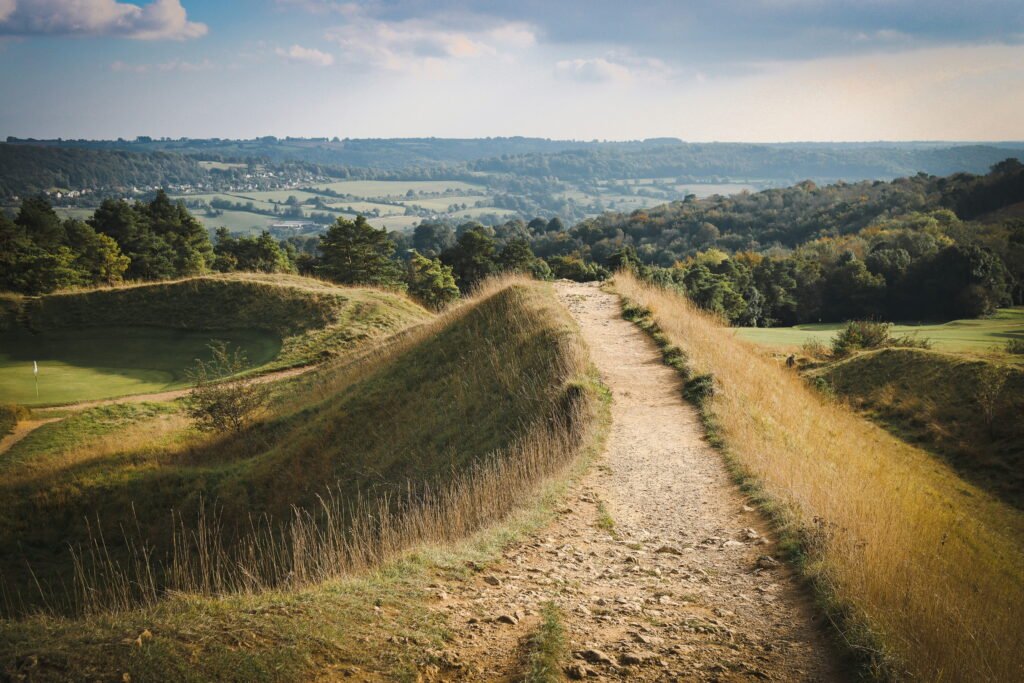Rocky walking trail following the top of a grassy ridge with sweeping views across the Cotswolds countryside. Wooded hills patchwork fields and a distant village stretch across the valley beneath a partly cloudy sky.